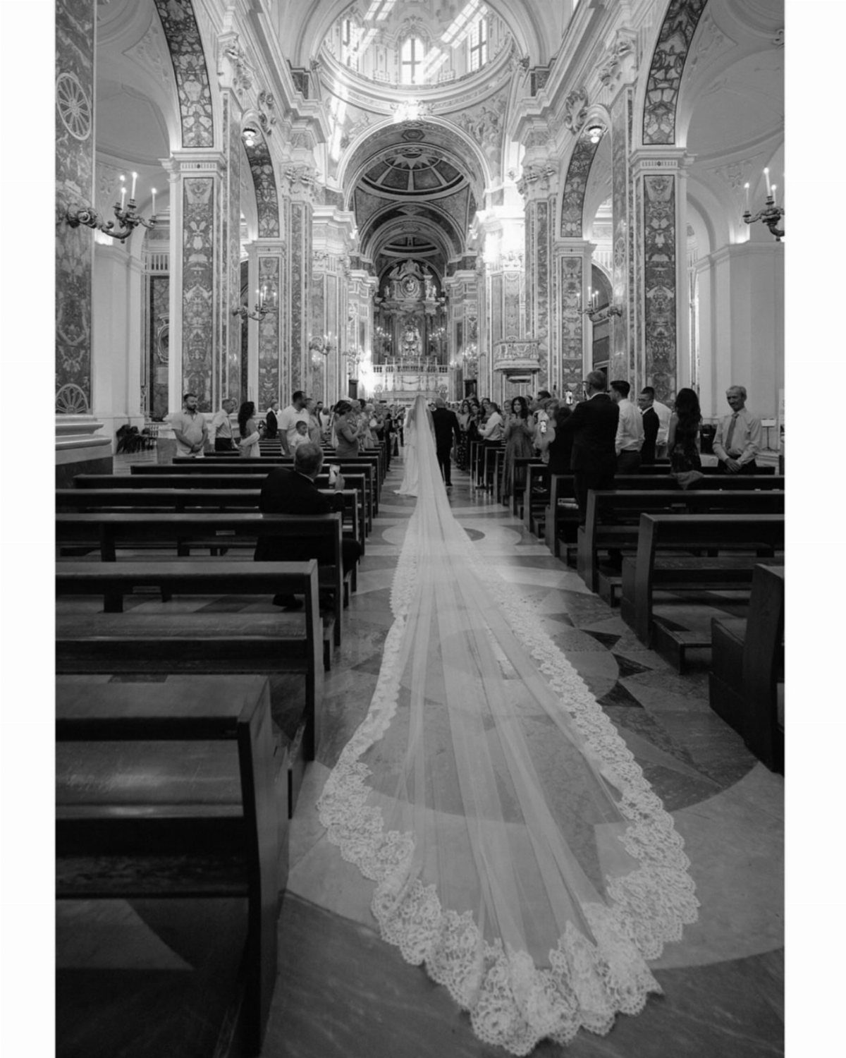 Bride walking down cathedral aisle with a very long lace-edged wedding veil trailing behind.