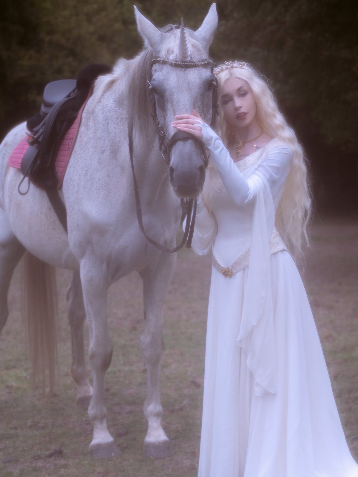 Woman in a white medieval wedding dress standing beside a white horse outdoors.