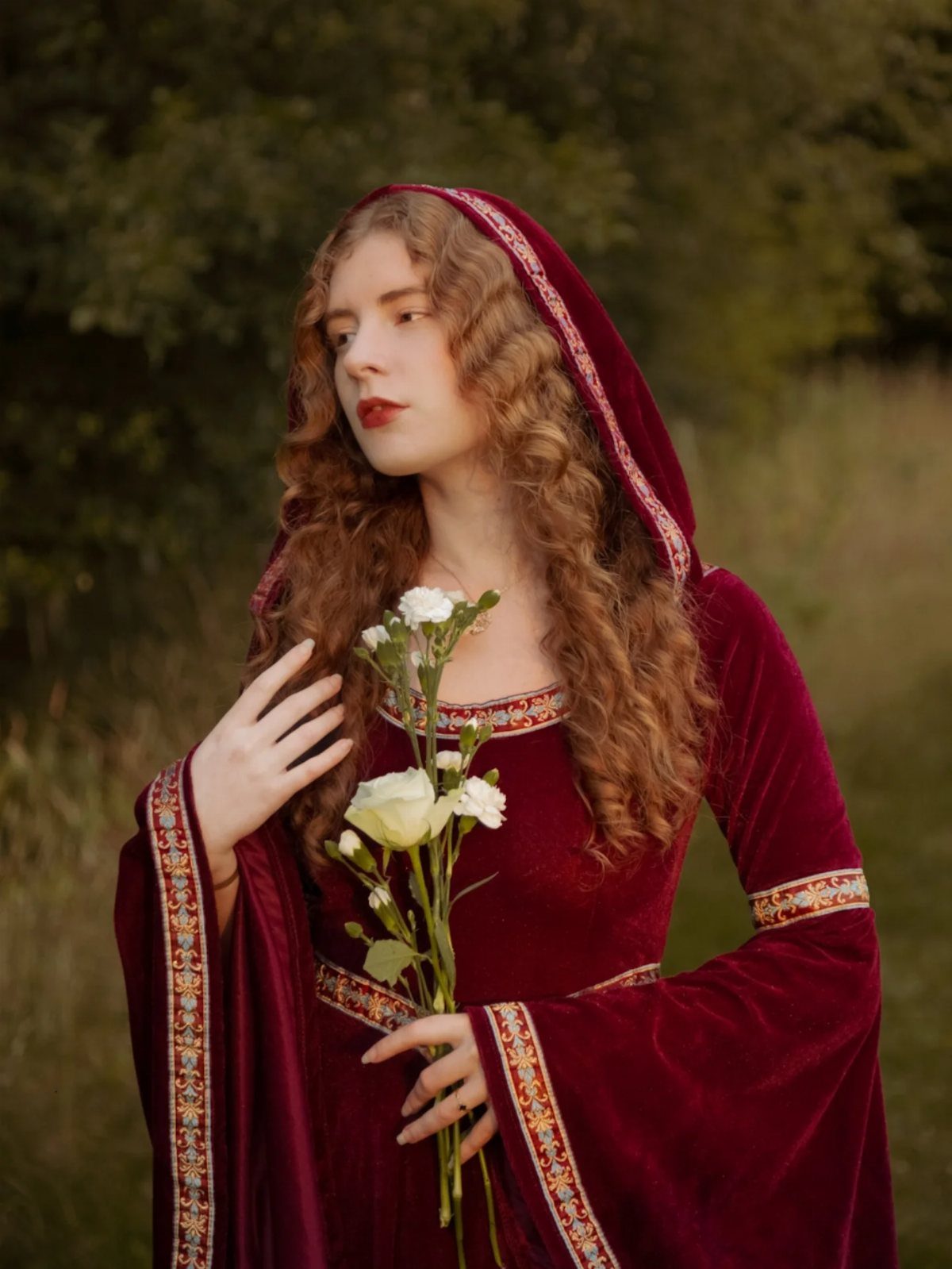 Woman in red velvet medieval gown with hood holding white roses outdoors