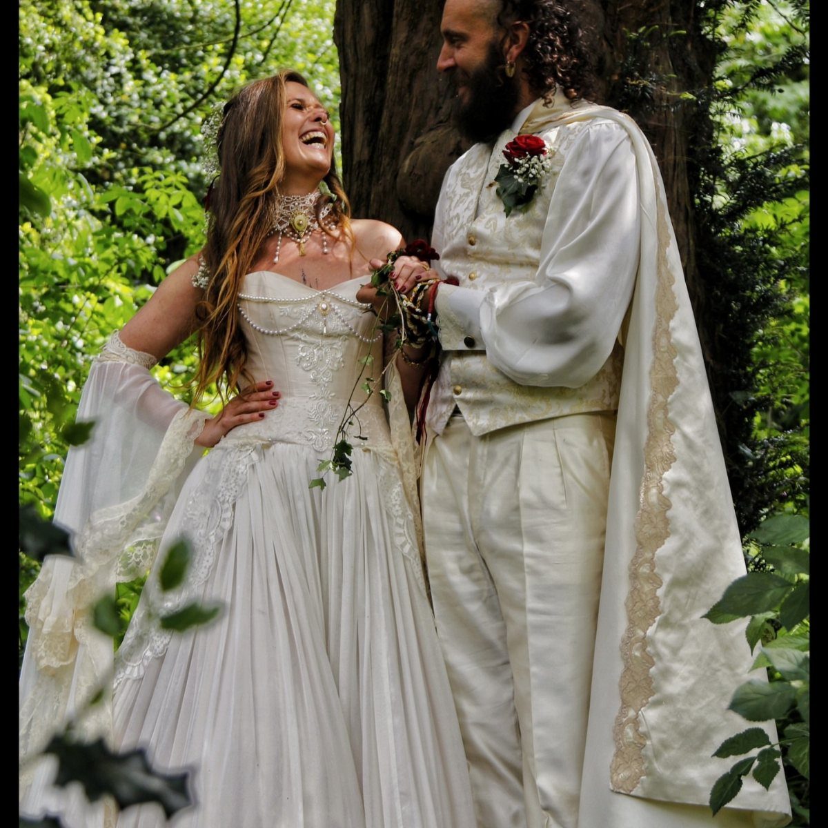 Couple in medieval-inspired wedding attire laughing together in a lush green forest