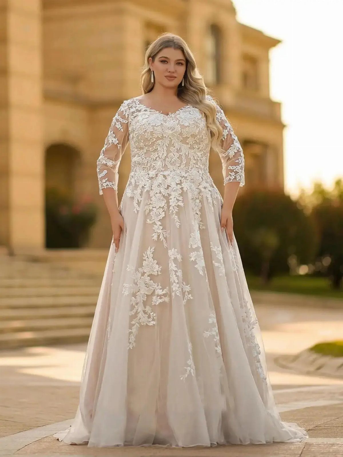 Curvy woman in a white lace wedding dress standing before a stone building.