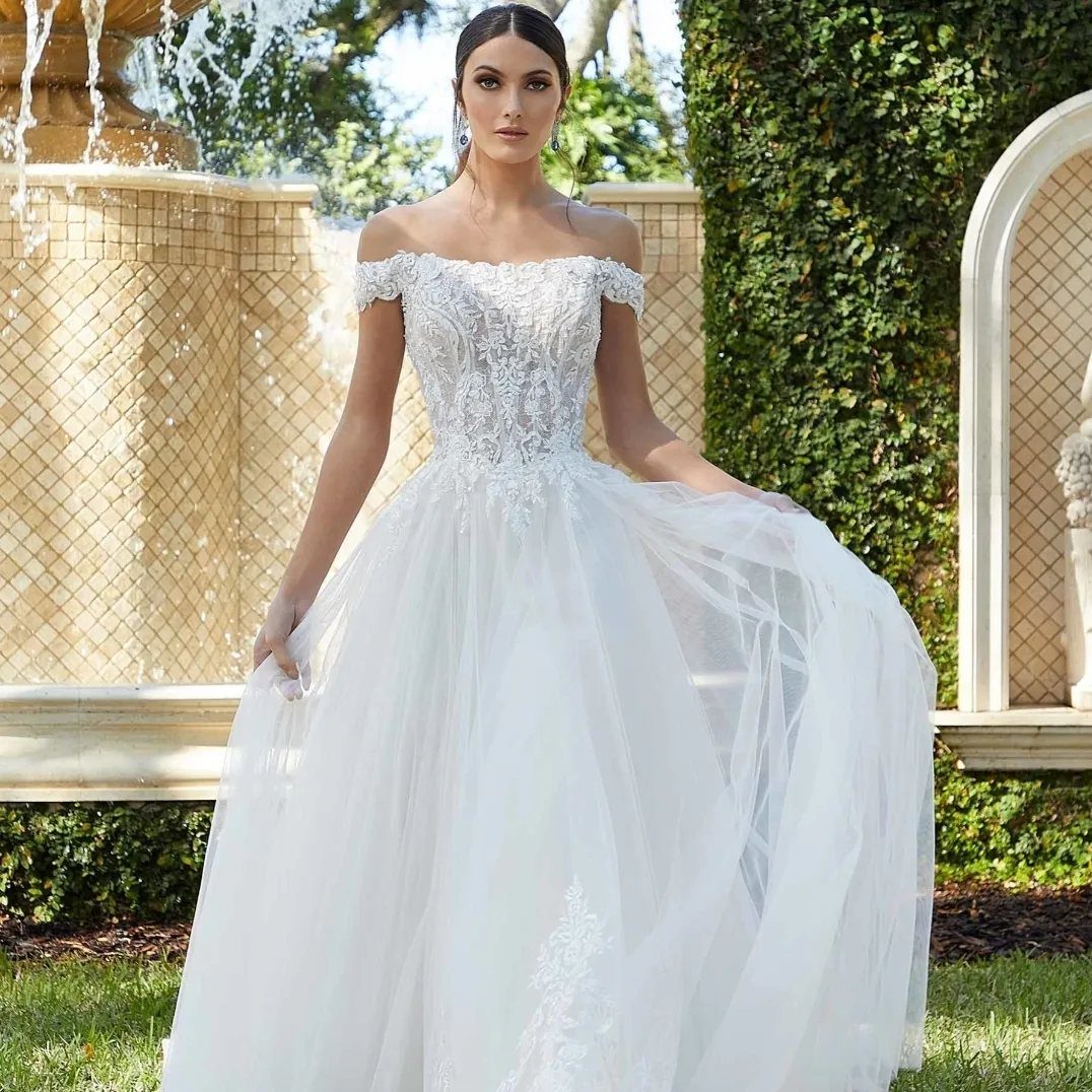 Woman in a white off-the-shoulder lace wedding dress standing by a stone fountain.