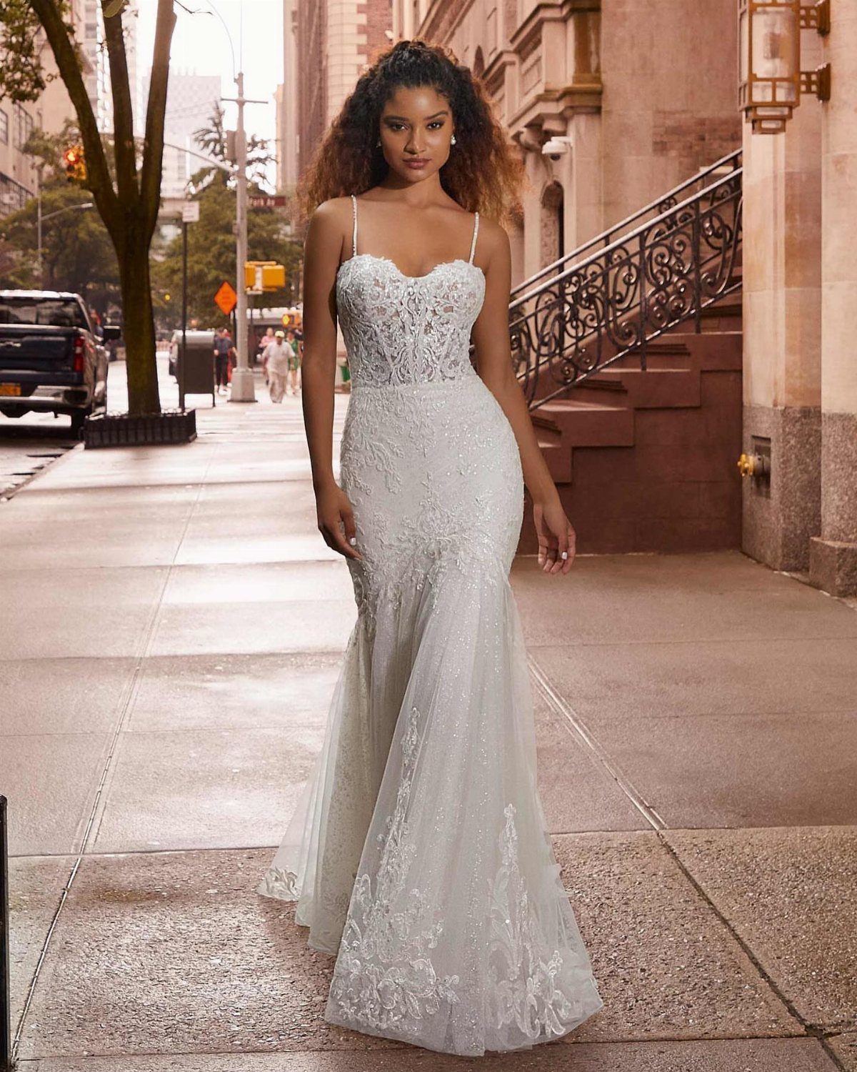Woman wearing a white lace floor-length wedding dress on a city sidewalk.