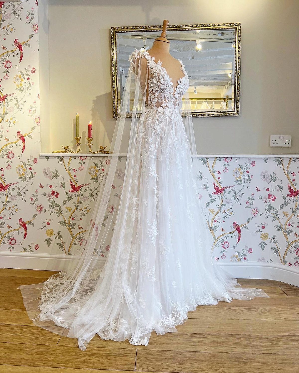 White lace wedding gown with a sheer cape on a mannequin against floral wallpaper.