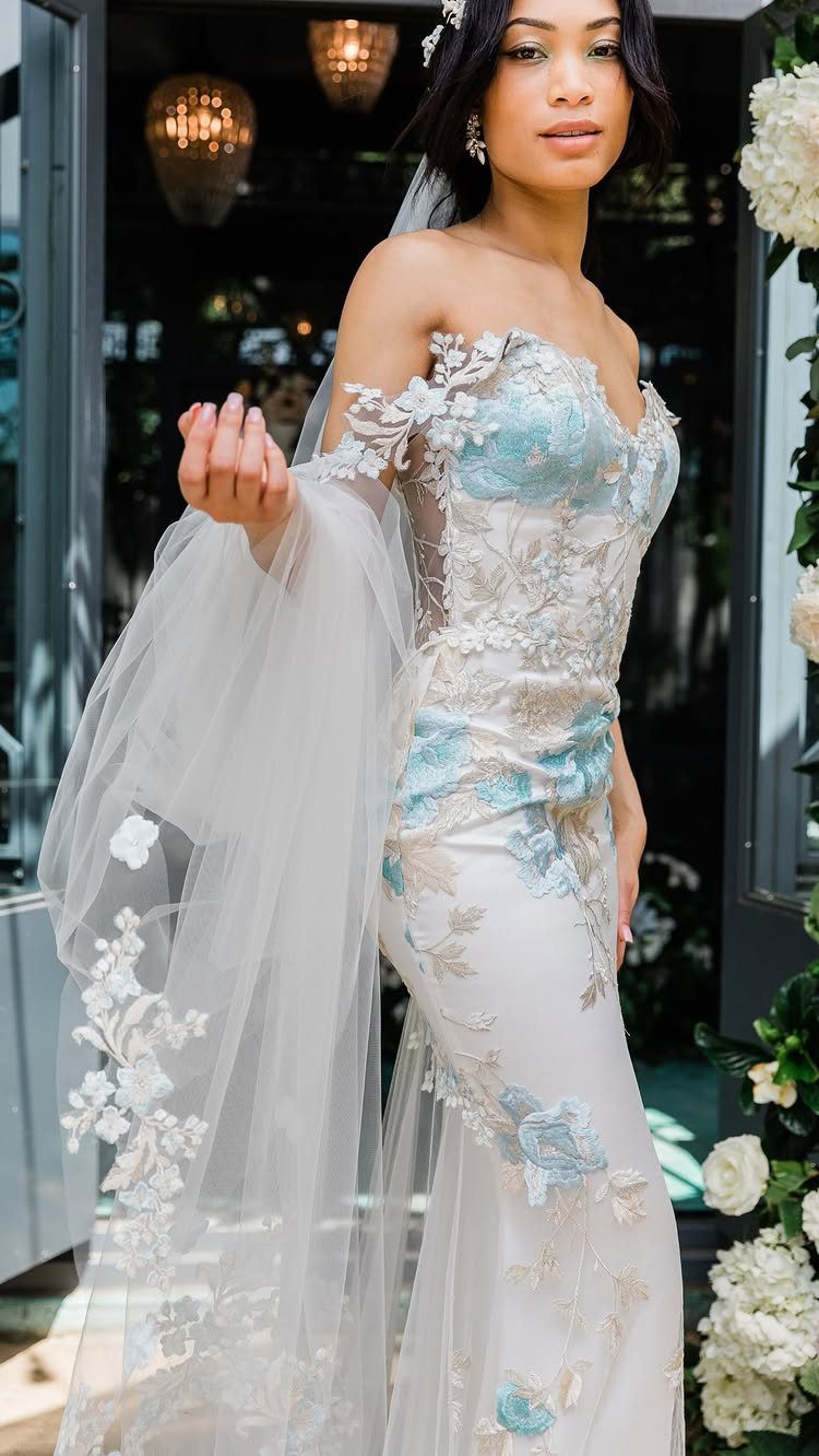 Woman in a white wedding dress with blue floral embroidery holding a sheer veil.