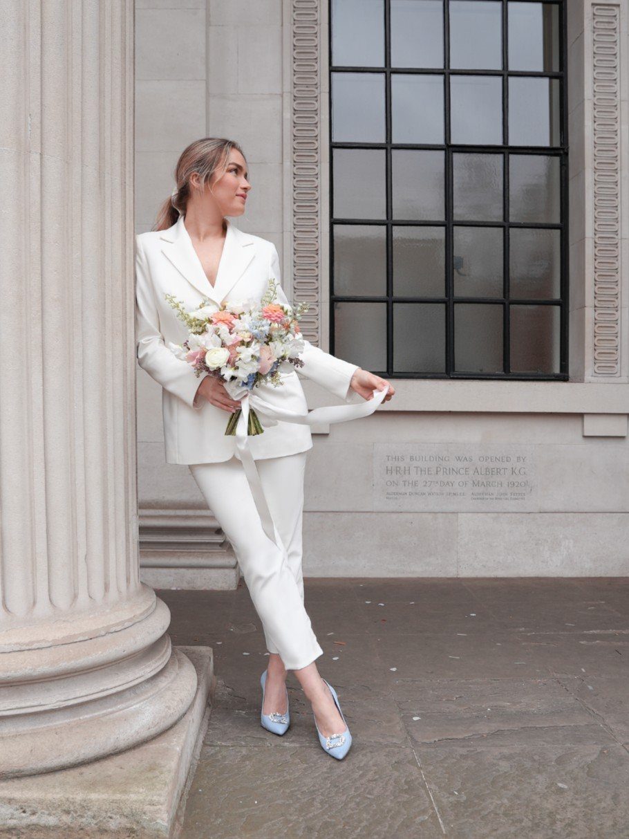 Woman in white bridal suit wearing light blue pointed-toe wedding shoes and bouquet.
