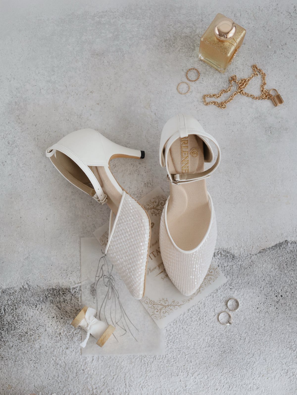 White pointed-toe bridal heels with ankle straps and gold wedding accessories on stone.