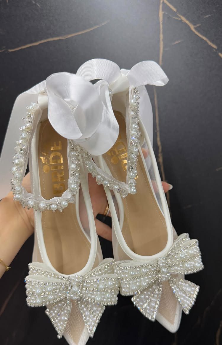 White pointed-toe bridal heels with pearl bows and satin ankle ribbons on dark marble.