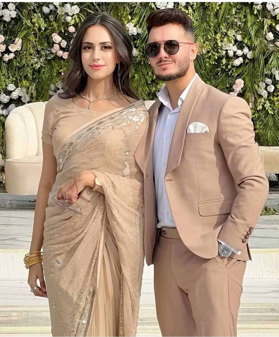 Man in tan suit and woman in beige saree posing before floral wall