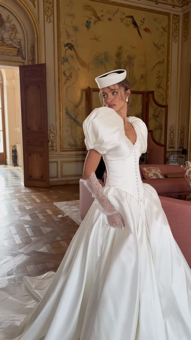 Woman in a white satin wedding gown with dramatic puffed sleeves and pillbox hat.