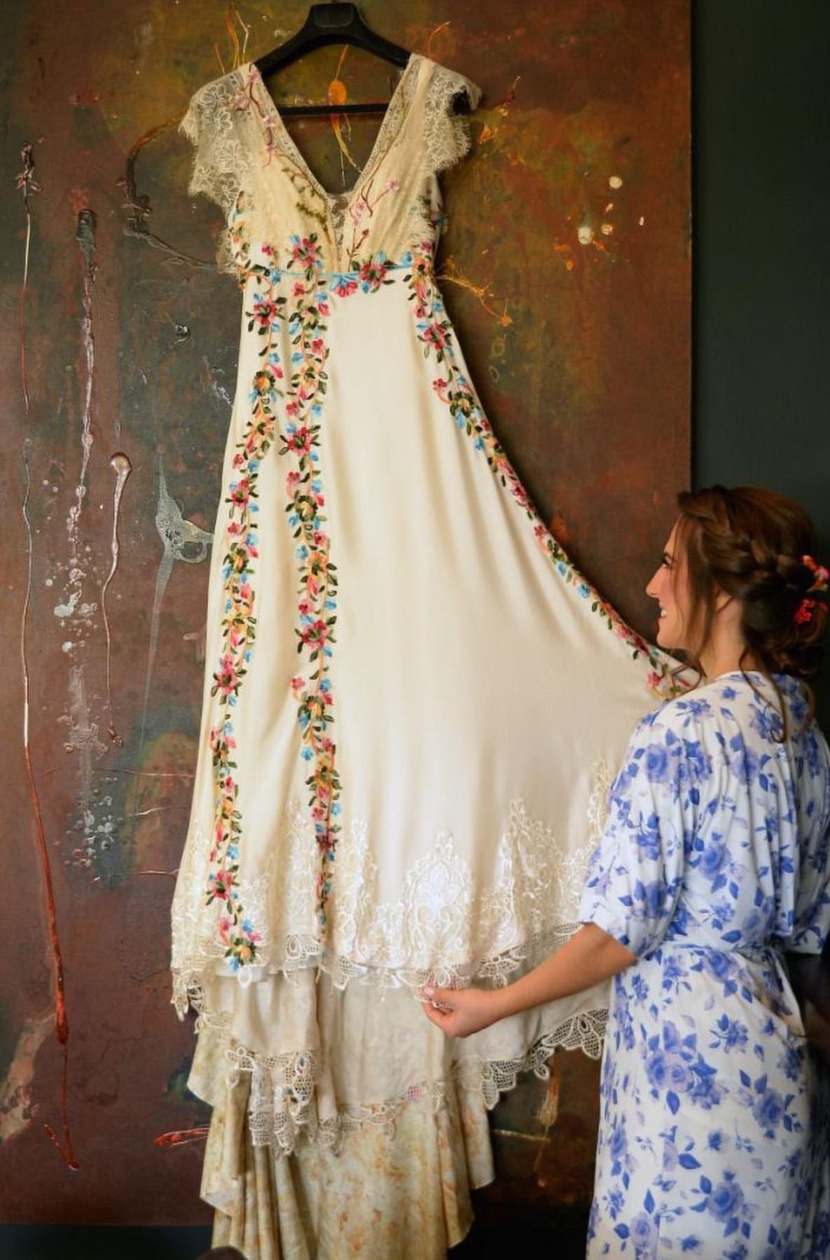 Cream wedding gown with colorful floral embroidery hanging against a textured brown background.
