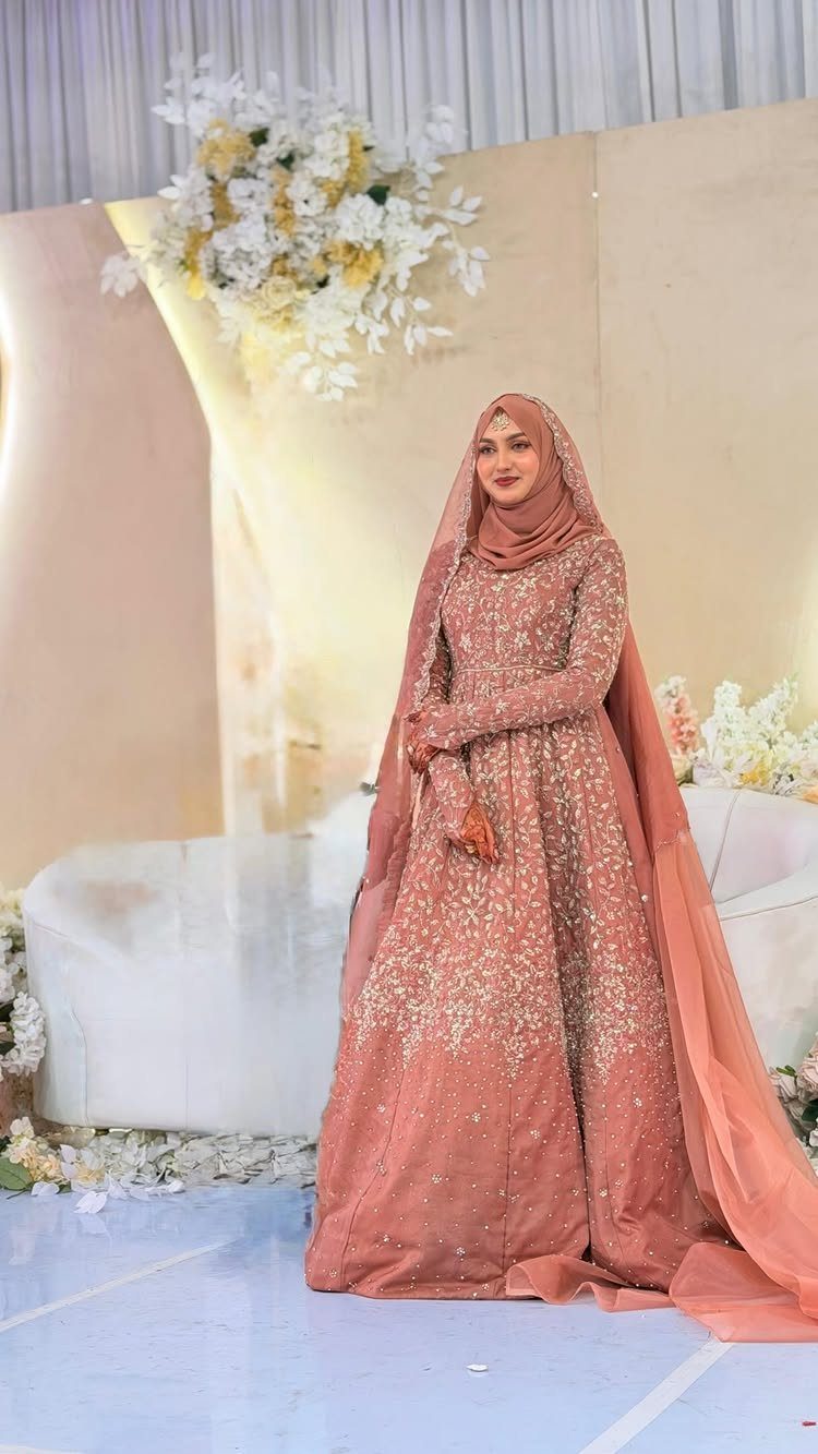 Woman in a dusty rose beaded gown and matching hijab standing by flowers.