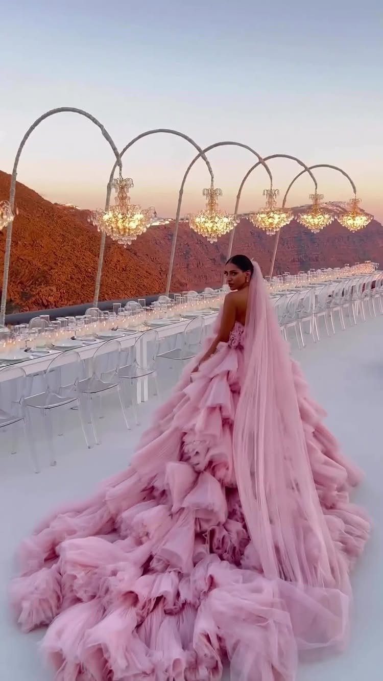 Woman in a voluminous ruffled pink wedding dress and veil standing outdoors at sunset.