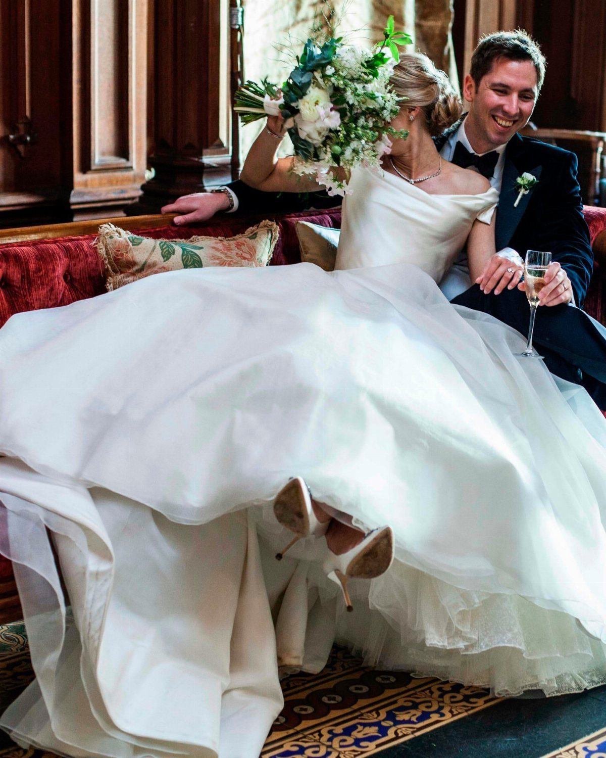 Bride in white organza gown and groom in tuxedo sitting on red sofa