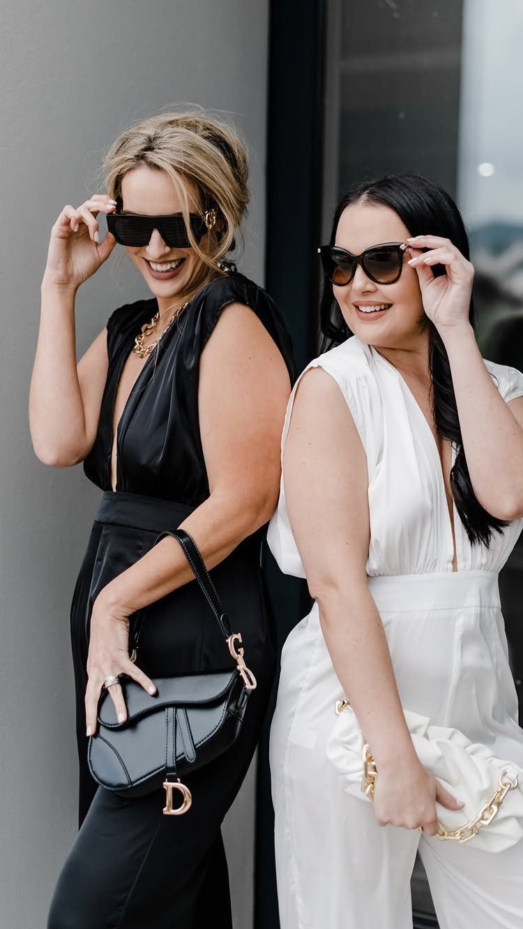 Two women wearing black and white sleeveless jumpsuits and sunglasses posing together.