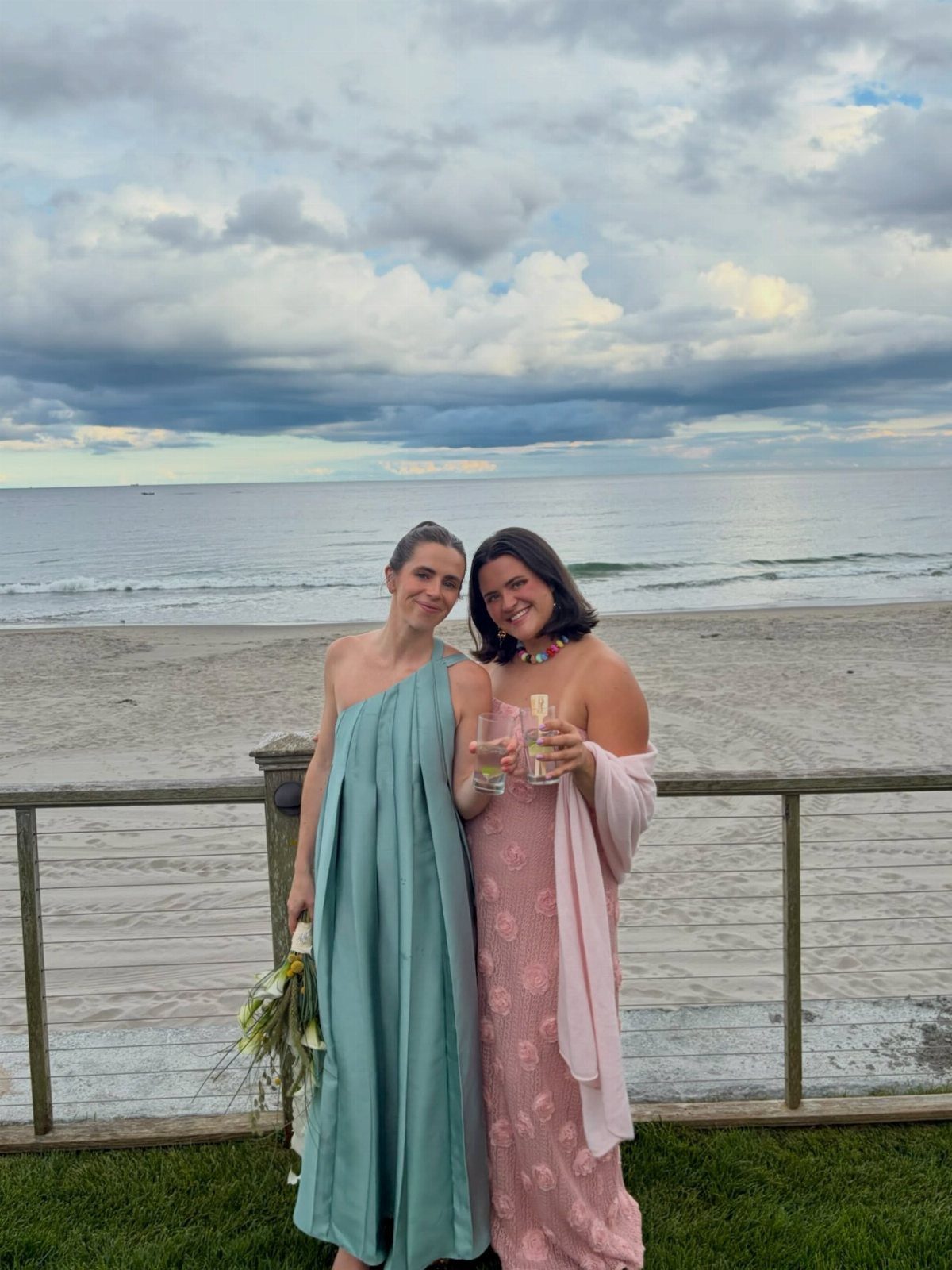 Two women in flowing blue and pink dresses posing on a beach deck.