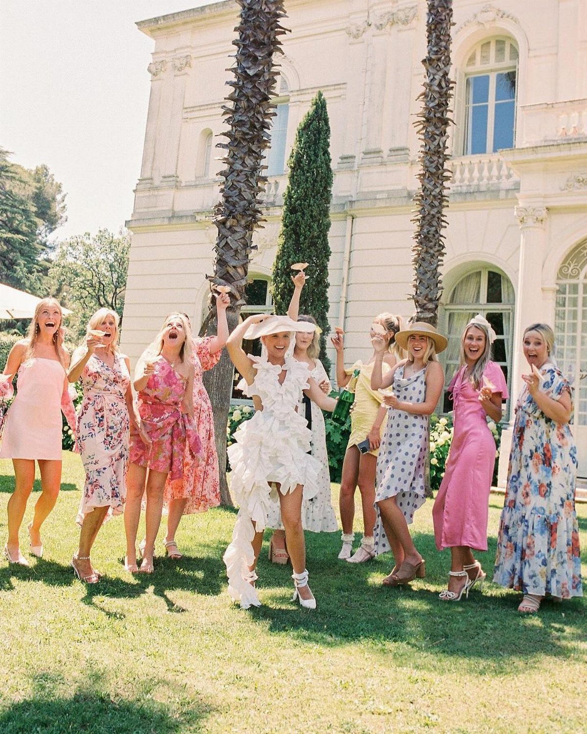 Women in colorful summer dresses laughing on a lawn before a stone villa.