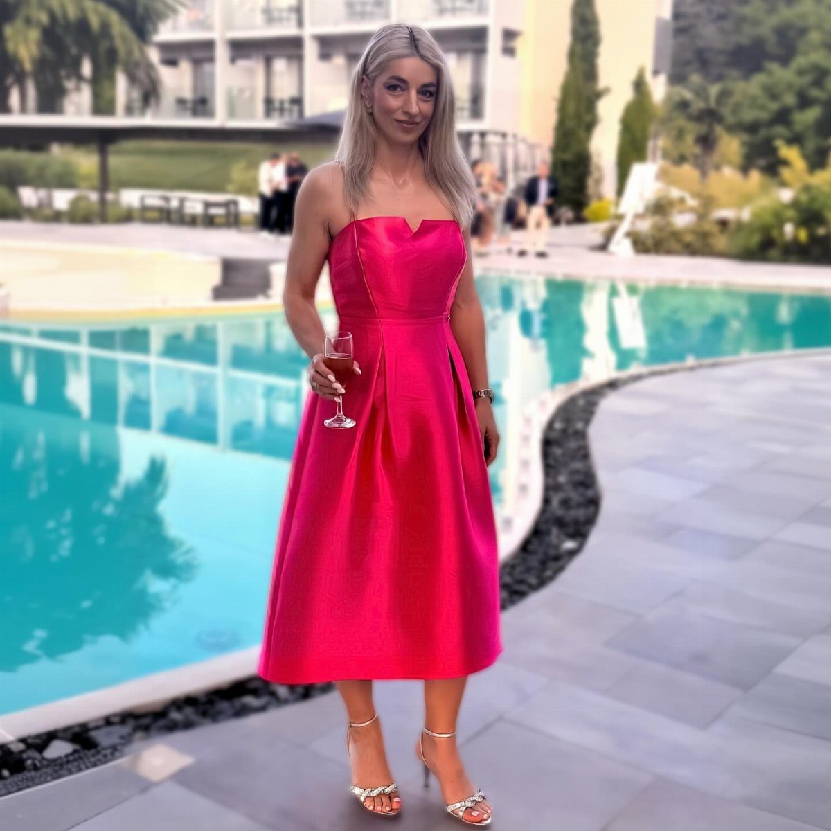 Woman in a bright pink strapless midi dress holding wine by a pool