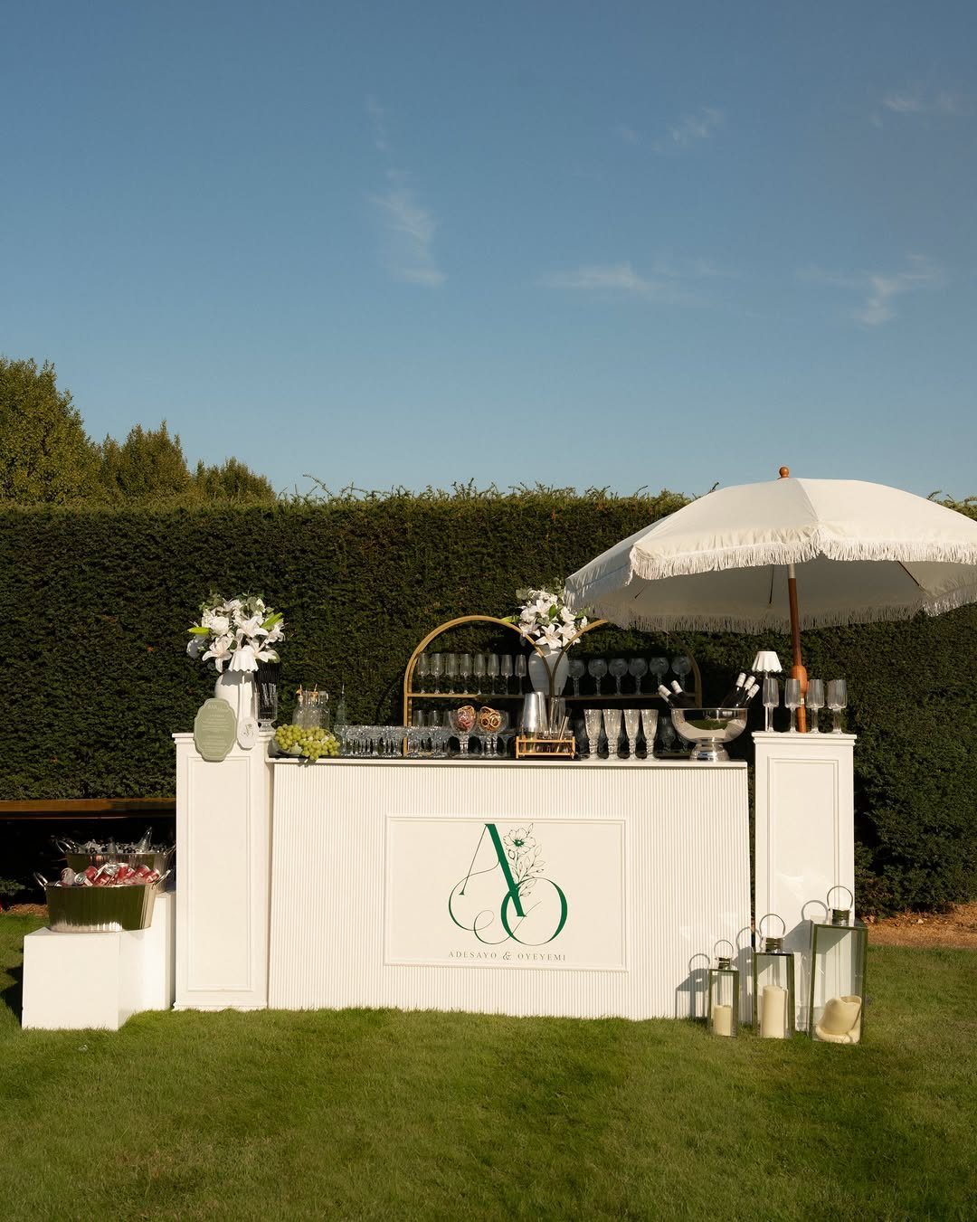 White outdoor bar station with glassware and umbrella on a green lawn