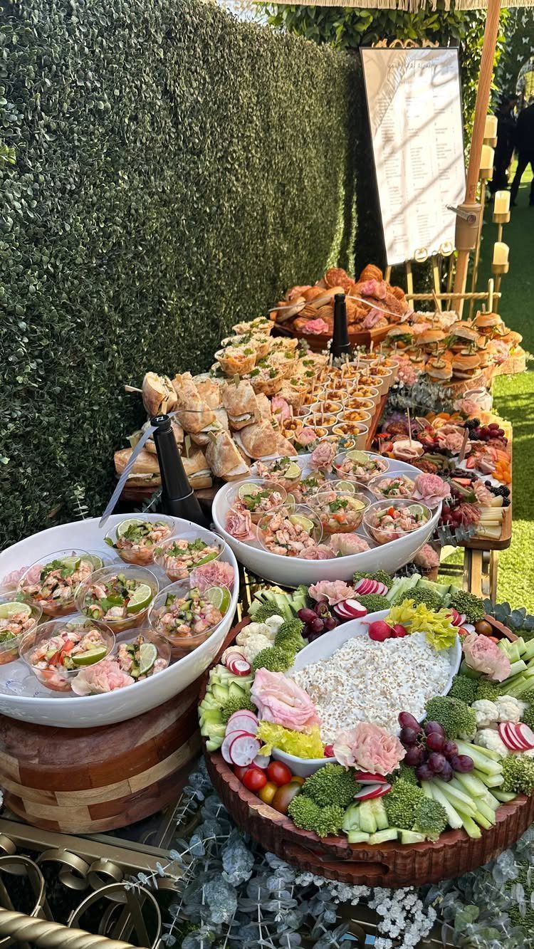 Lavish food table with shrimp cocktail, vegetable platters, and mini sandwiches outdoors.