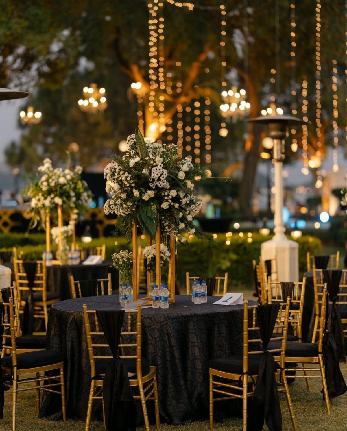 Round wedding table with black linens, gold chairs, and tall white floral centerpieces.