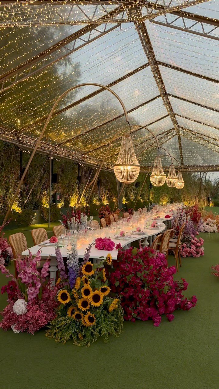 Curved white wedding table decorated with vibrant flowers under a lit transparent tent.