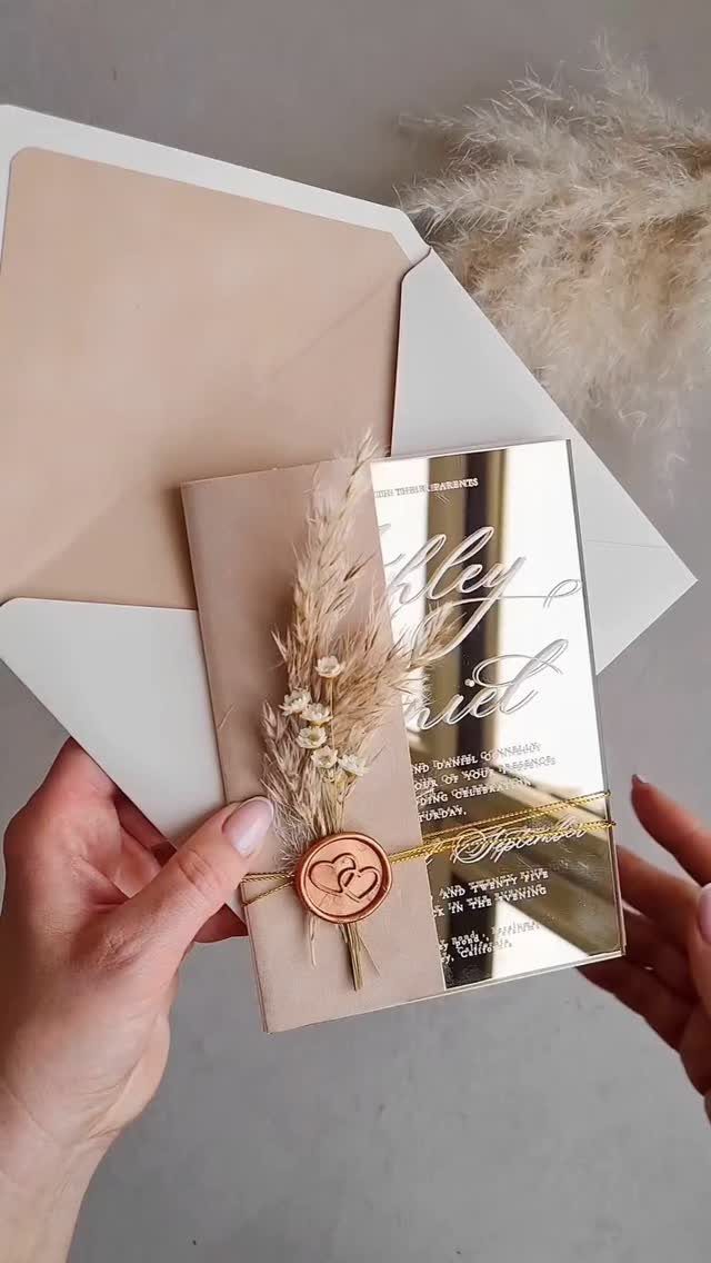 Wedding invitation suite with mirrored acrylic card, beige envelope, and dried floral accents.