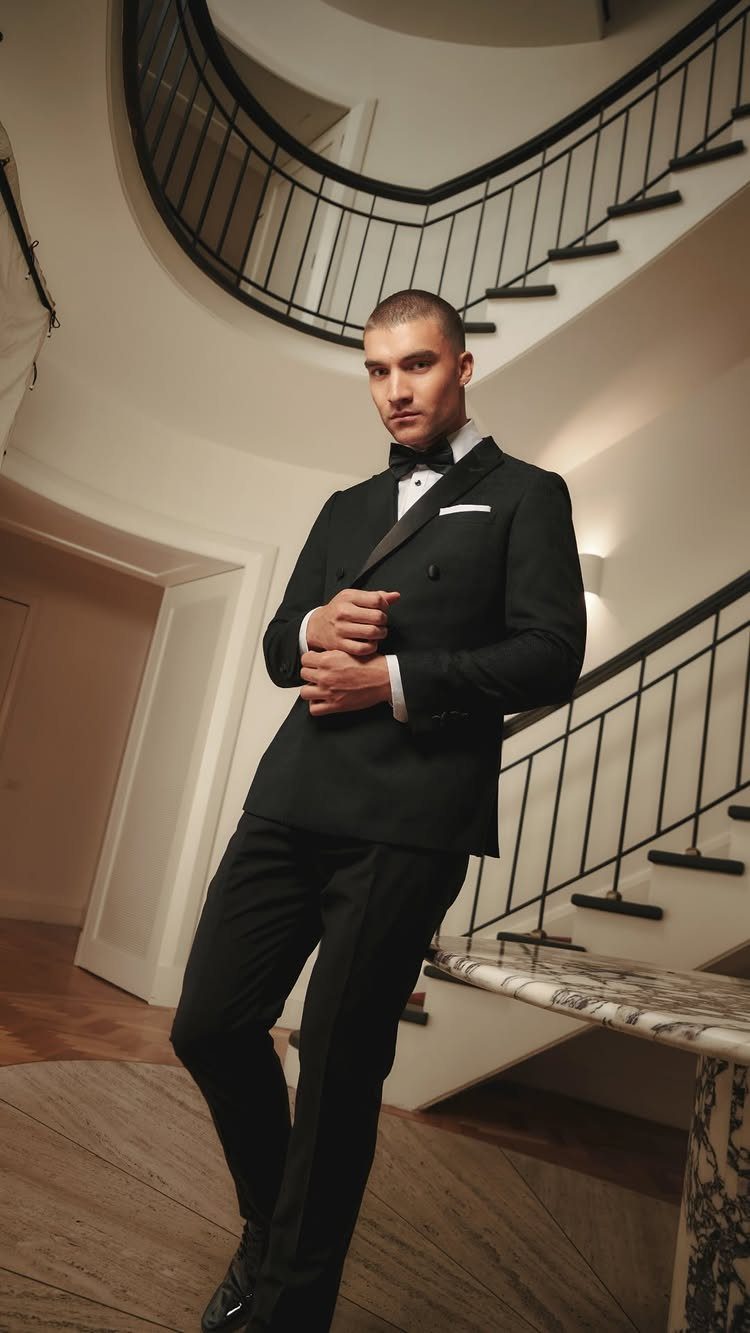 Man in a black tuxedo and bow tie posing by a curved staircase.