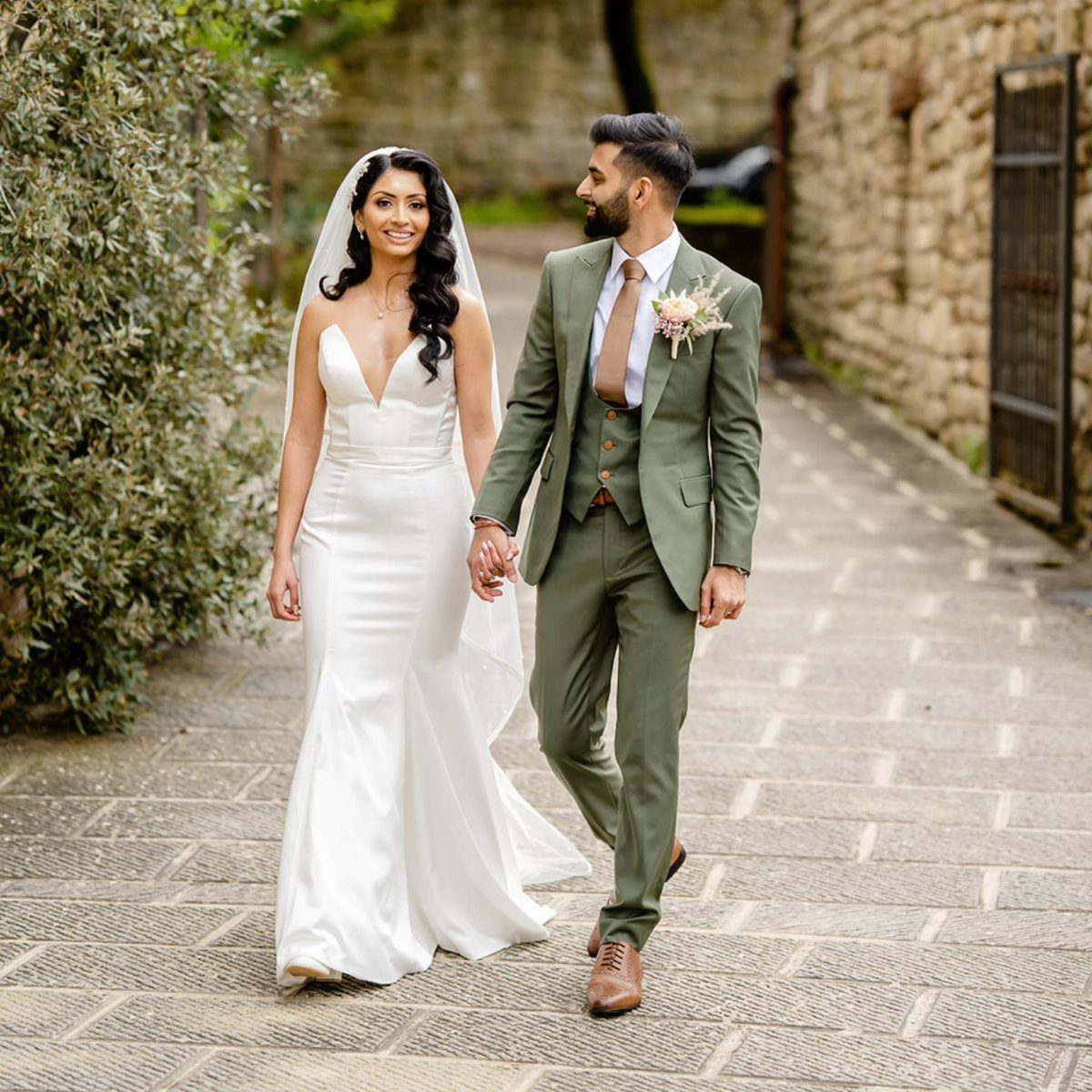 Bride in white dress and groom in olive green suit walking together outdoors.
