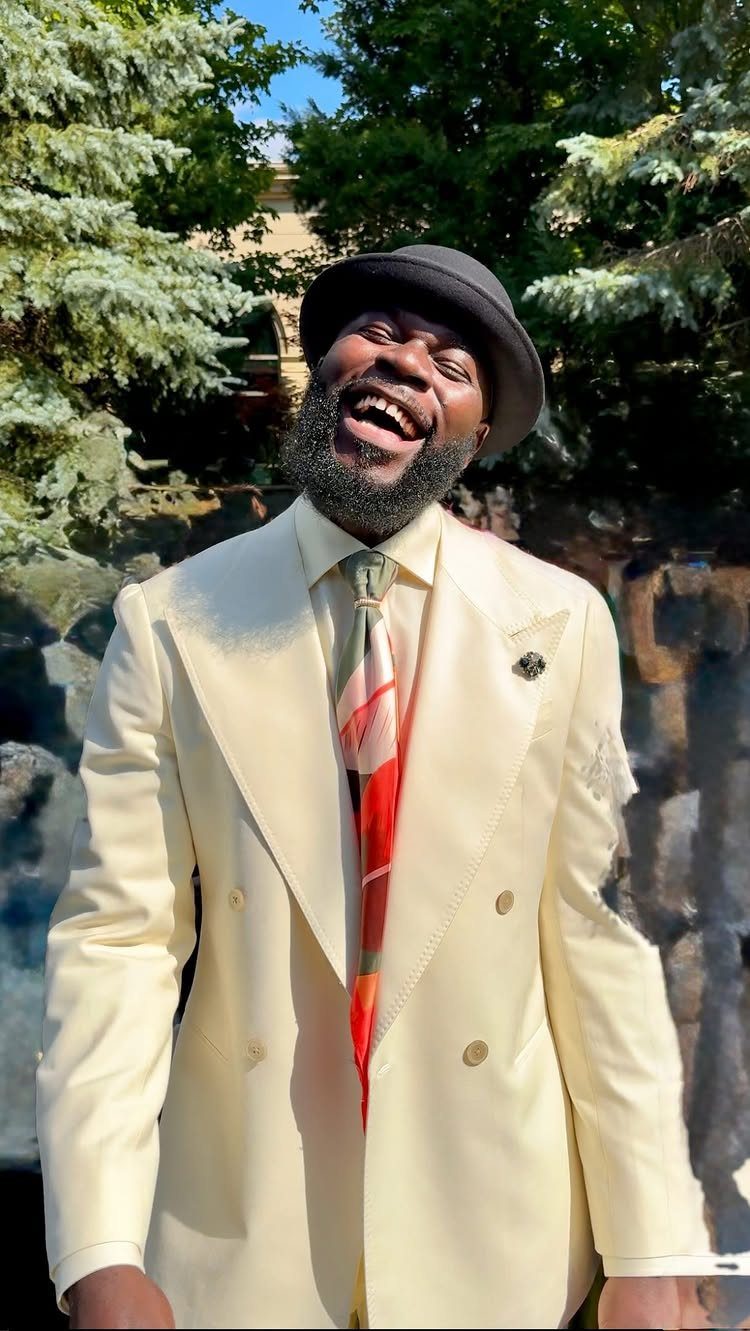 Man in a cream double-breasted suit and black bowler hat smiling outdoors.