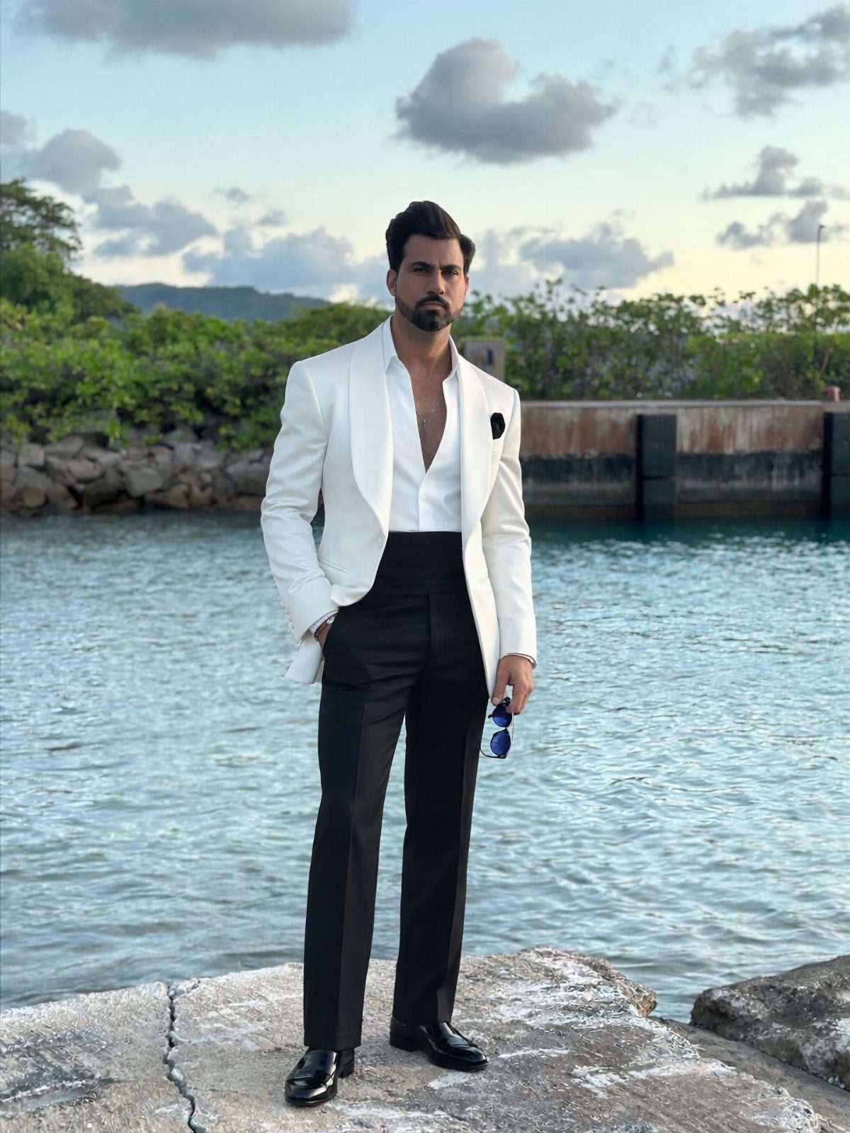 Man in a white tuxedo jacket and black trousers standing by the ocean.