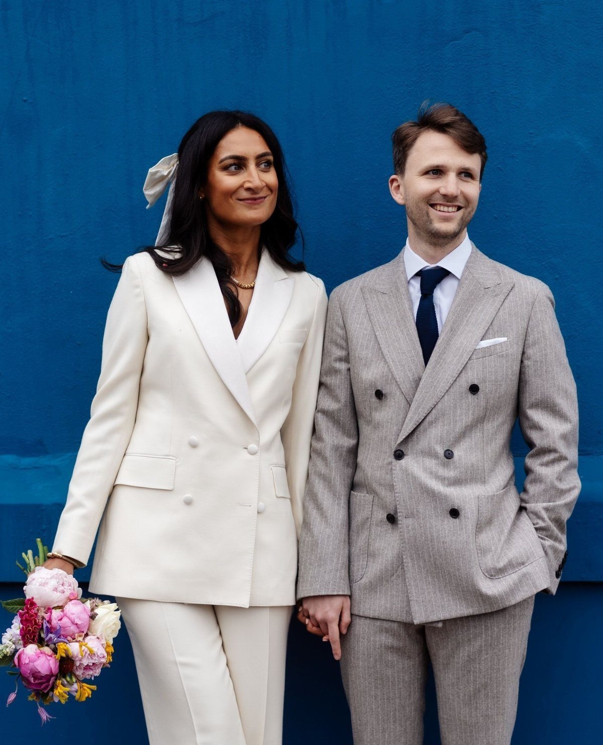 Woman in white bridal suit and man in grey suit against blue wall