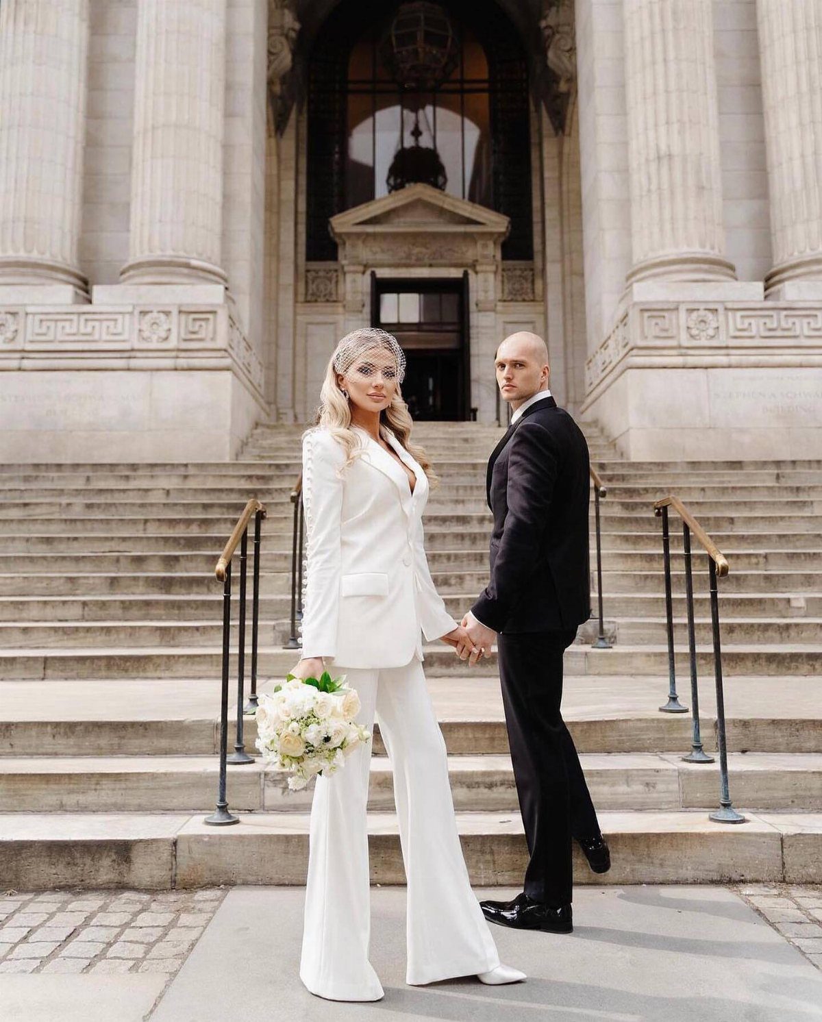 Woman in white bridal pant suit holding bouquet standing with groom outdoors