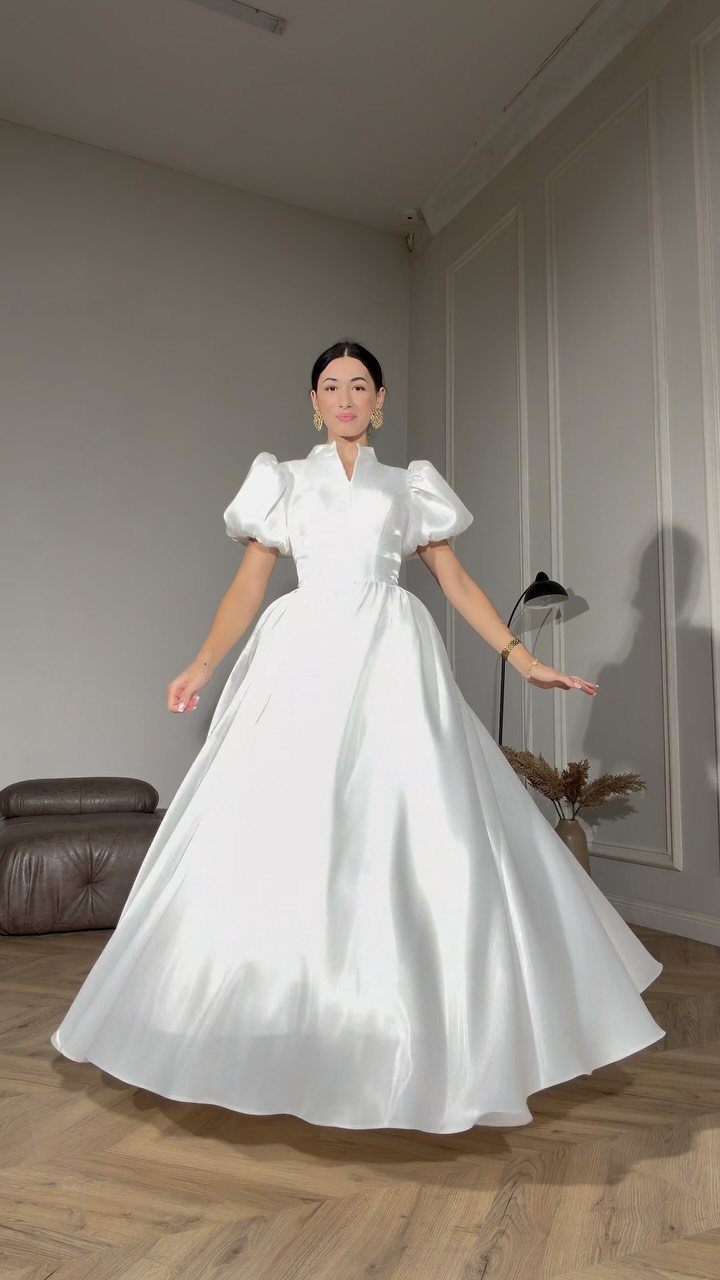 Woman wearing a white satin wedding dress with puff sleeves in a room.