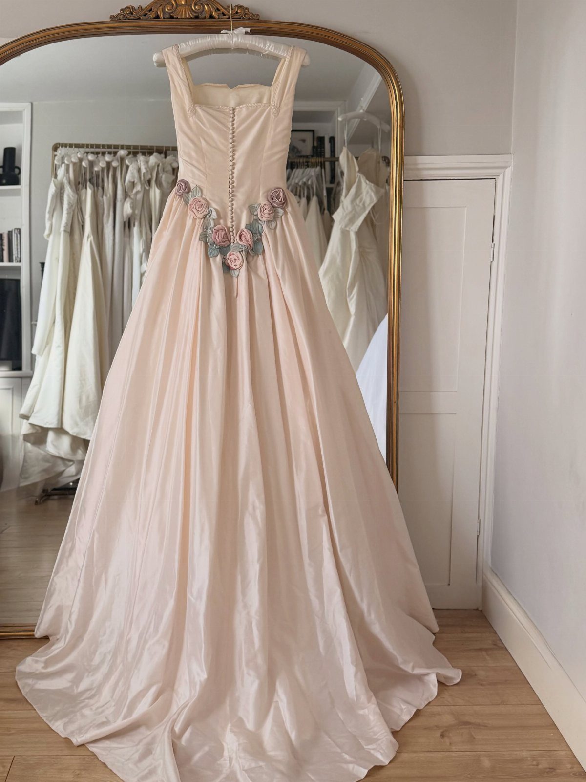 Blush wedding gown with floral waist details hanging before a gold mirror.