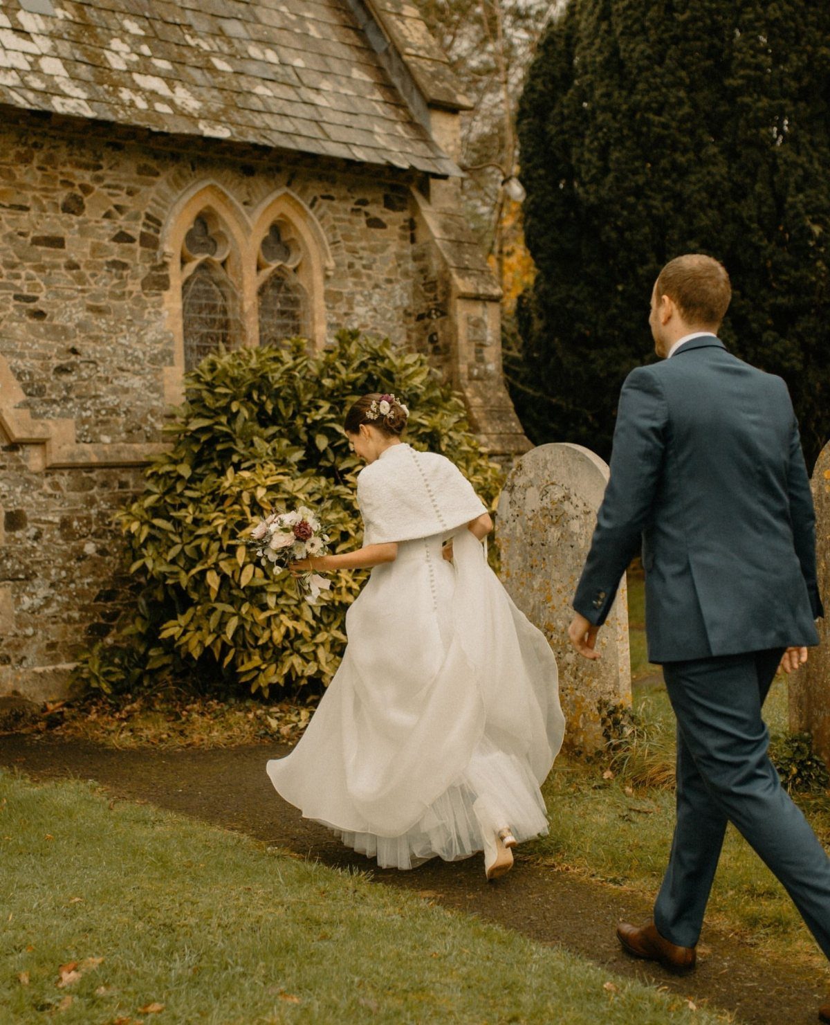 Bride in white wedding dress and capelet walking with groom near stone church