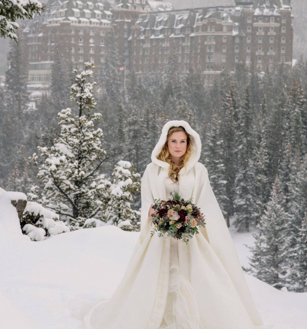 Bride in a white hooded cape and wedding dress holding flowers in snowy mountains