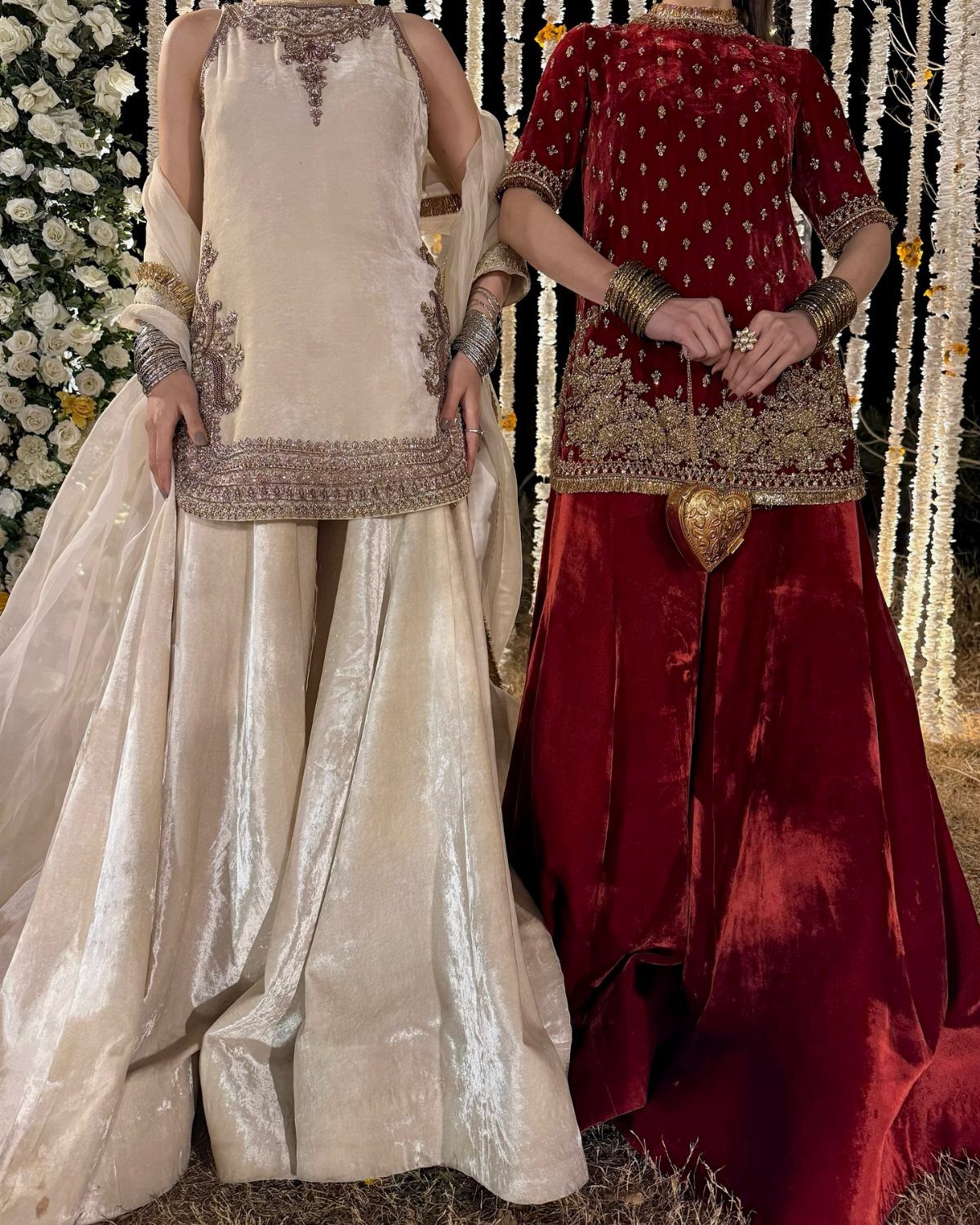 Two women wearing ornate cream and red velvet bridal dresses with gold embroidery.
